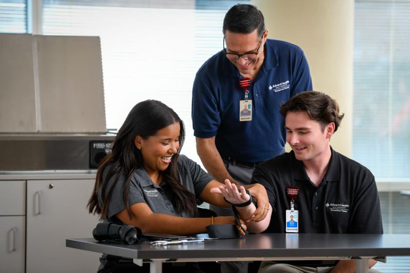 Image of faculty member smiling and helping two students with a hand therapy machine