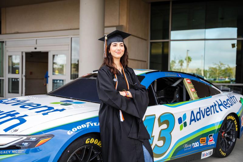 A student in cap and gown stands next to the AdventHealth Nascar.