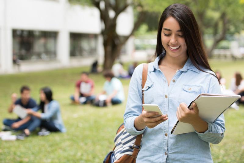 A female student walking outdoors holding her books and looking at her phone