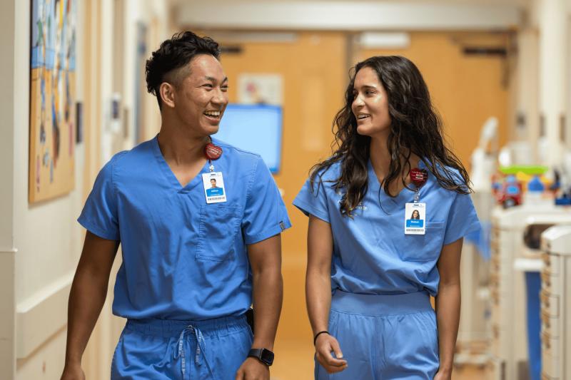 A young male and young female nurse walk down a hospital hallway together