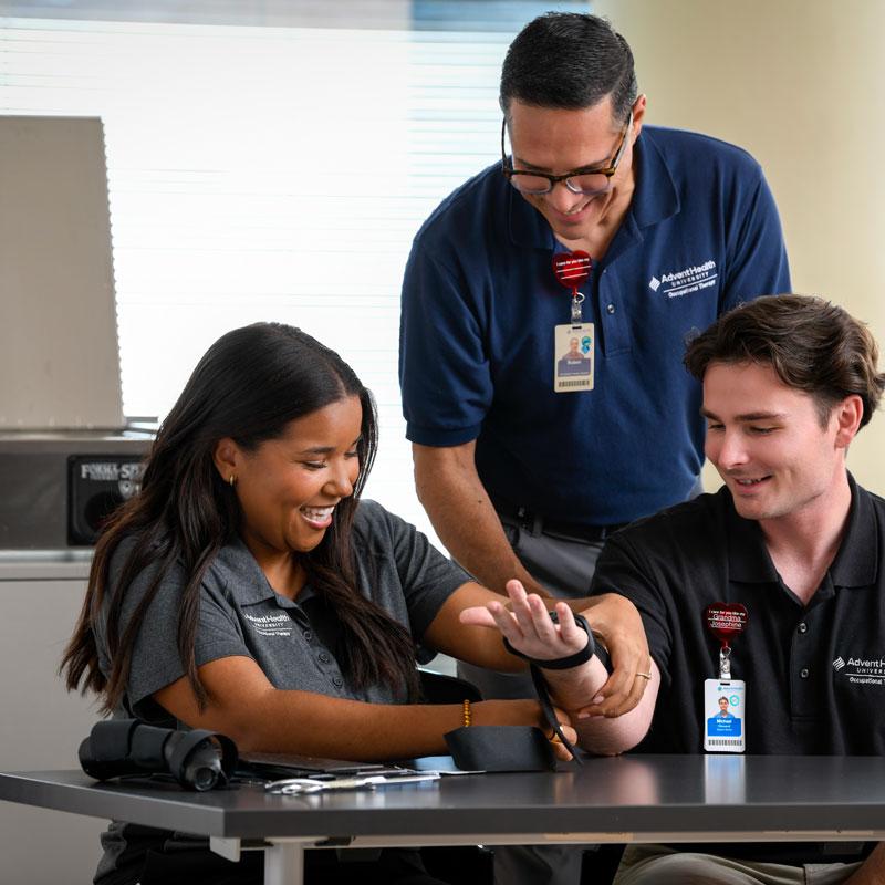 Image of faculty member smiling and helping two students with a hand therapy machine