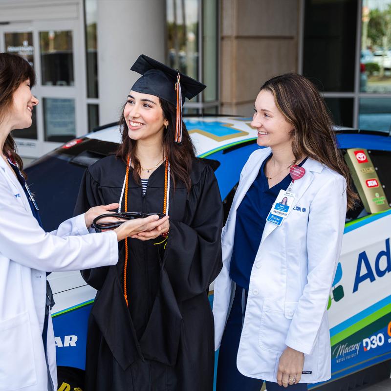 A graduate and two nurses standing in front of the AdventHealth Nascar