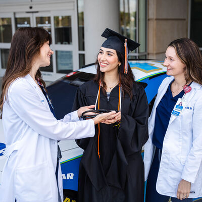 A graduate and two nurses standing in front of the AdventHealth Nascar