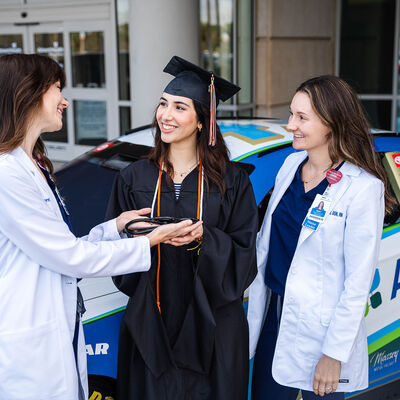 A graduate and two nurses standing in front of the AdventHealth Nascar