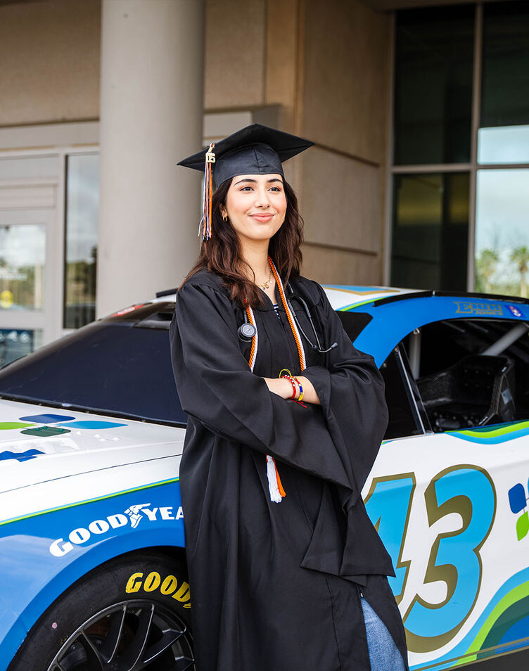 A student in cap and gown stands next to the AdventHealth Nascar.